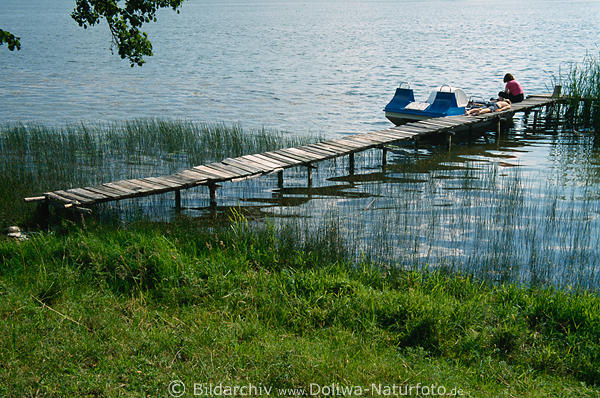 Seeufer-Steg mit Trettboot-Touristen liegen auf Holzsteg in Schilf Wasser Grnwiese Masuren Seeidylle
